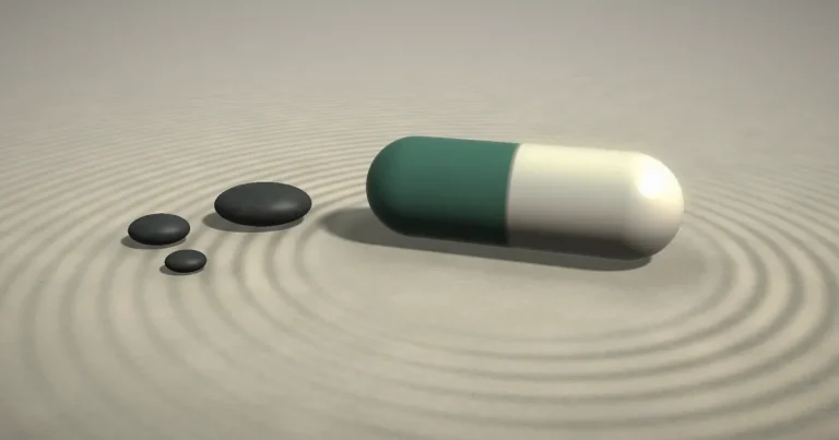 Two-tone sage and white capsule resting on raked sand with soft concentric ripples and smooth dark stones